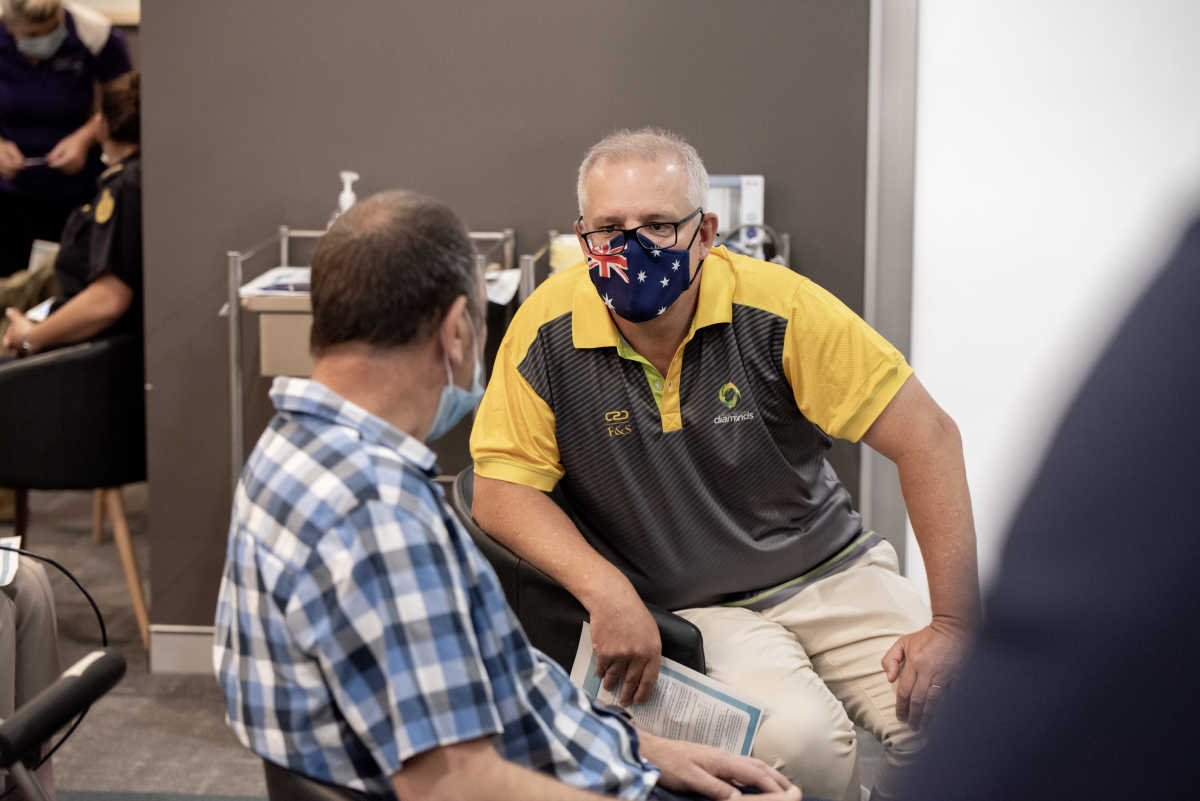 Life Without Barriers' client Brett with Prime Minister Scott Morrison talking before receiving the COVID-19 Vaccine. Brett has his back to the camera and is wearing a blue checked shirt. Prime Minister Morrison is wearing an Australian flag face mask and a yellow and grey t-shirt.