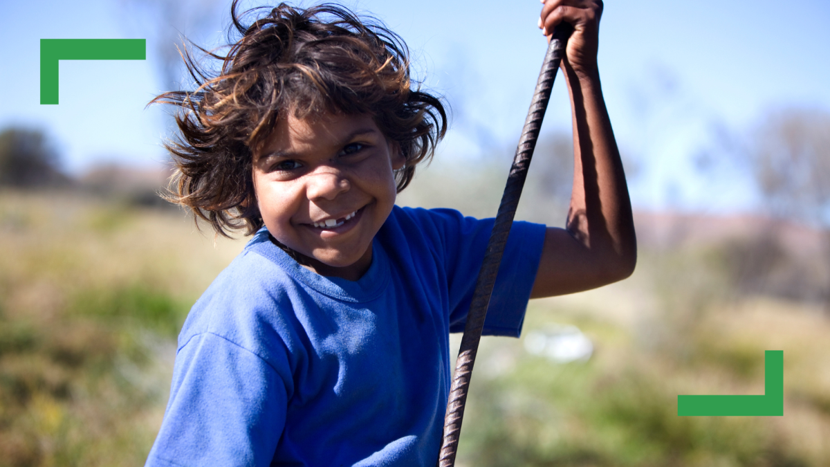 A young Aboriginal boy with brown hair is wearing a blue shirt and holding on to an iron pole, smiling at the camera.