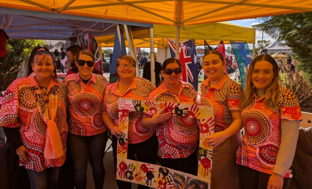 A group of staff wearing NAIDOC t-shirts stand together, holding up a cardboard cut-out decorated with handprints.
