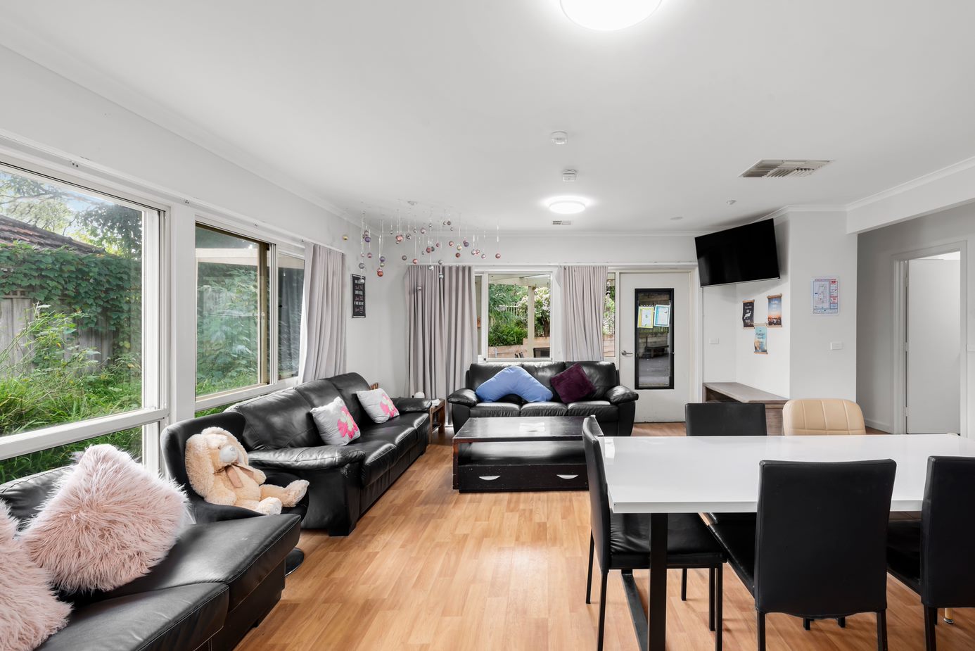 Lounge room with three black couches, a large coffee table, TV unit and a TV mounted to the wall. In the room is also a white dining table with six chairs.