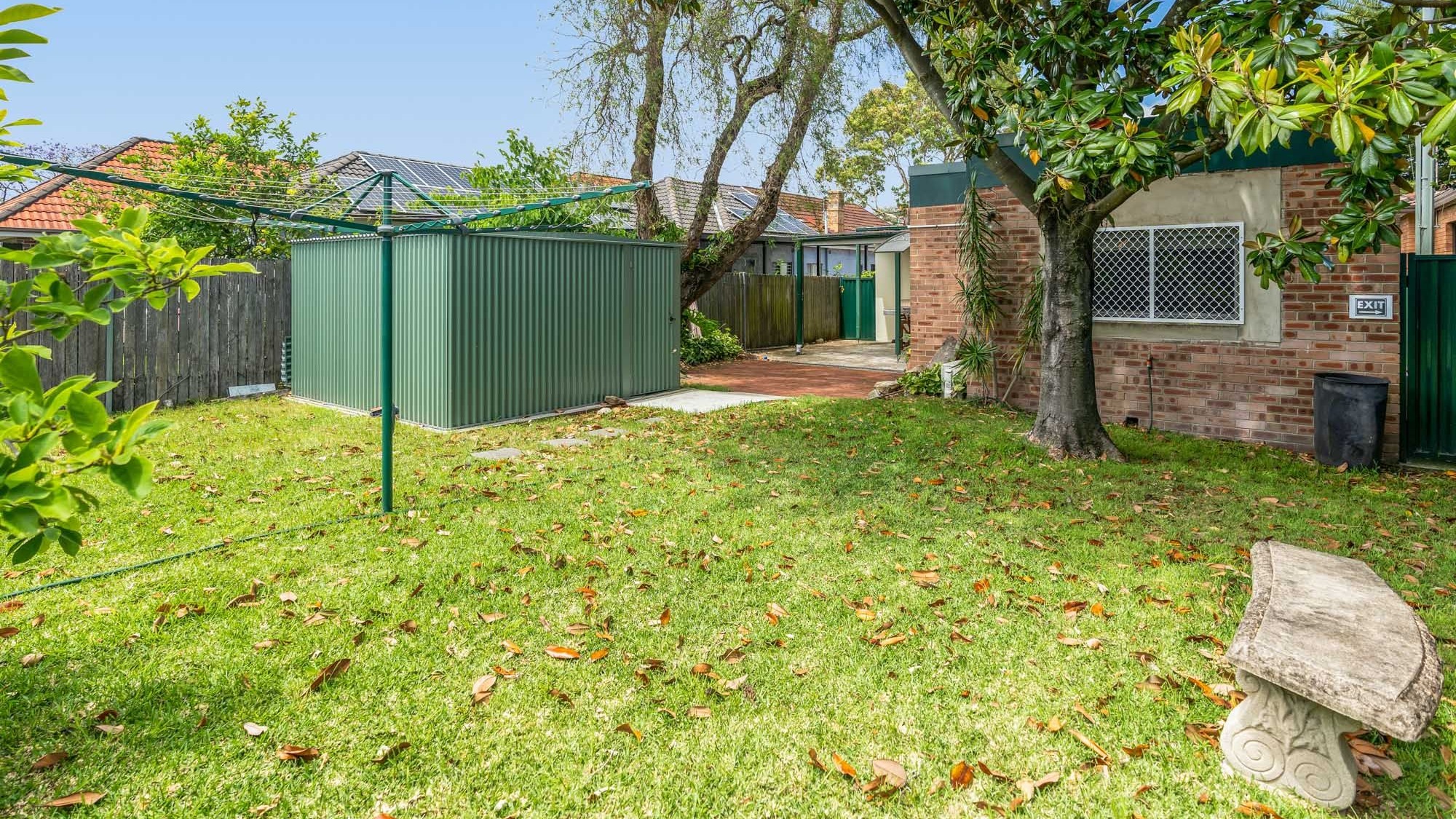 Spacious backyard with green grass, shady mature trees, clothes line and shed.