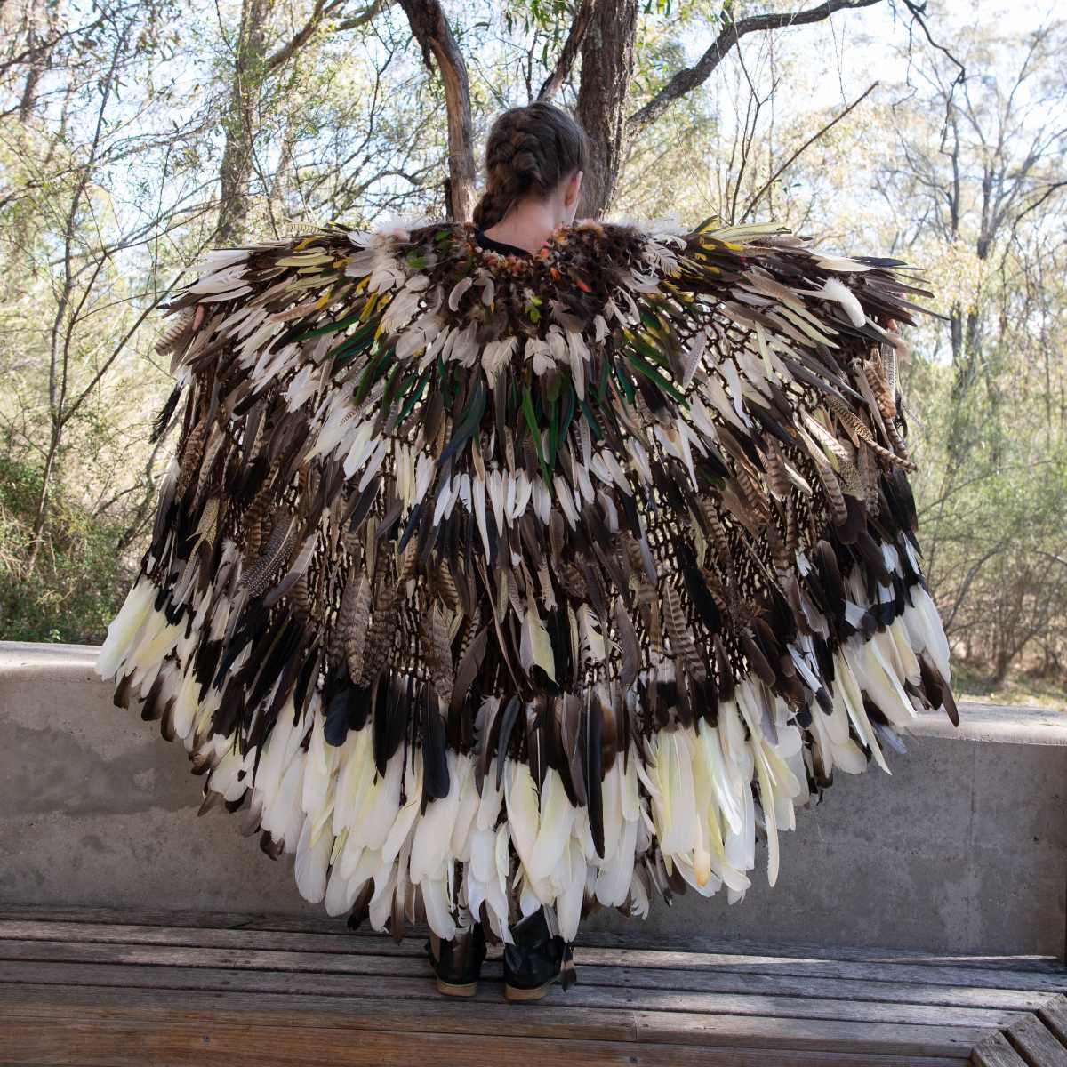A young girl is wearing a feather cloak and stands with her arms outstreched and her back to the camera