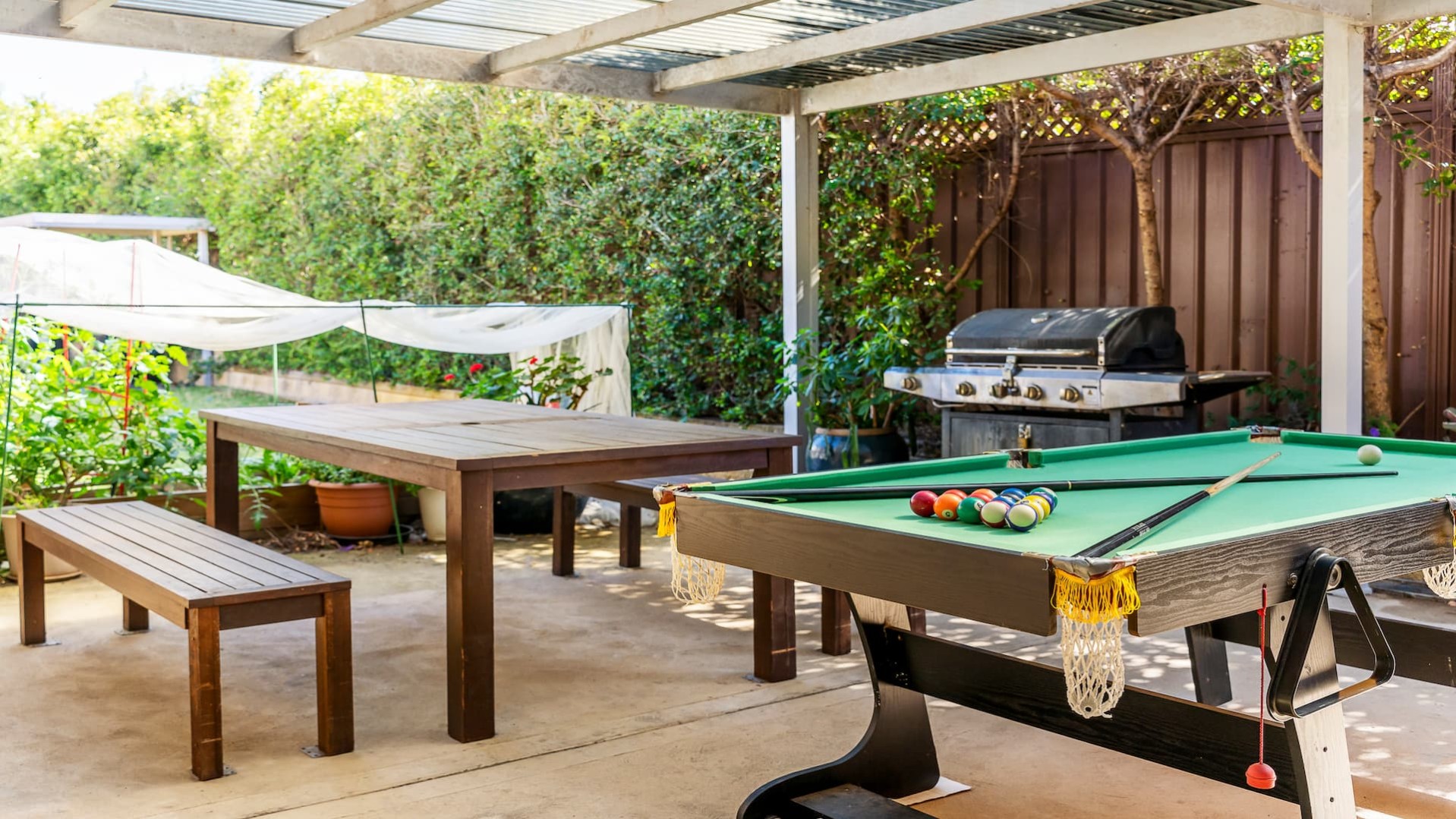 undercover patio with outdoor table and chairs, BBQ, pool table overlooking garden beds and hedge.