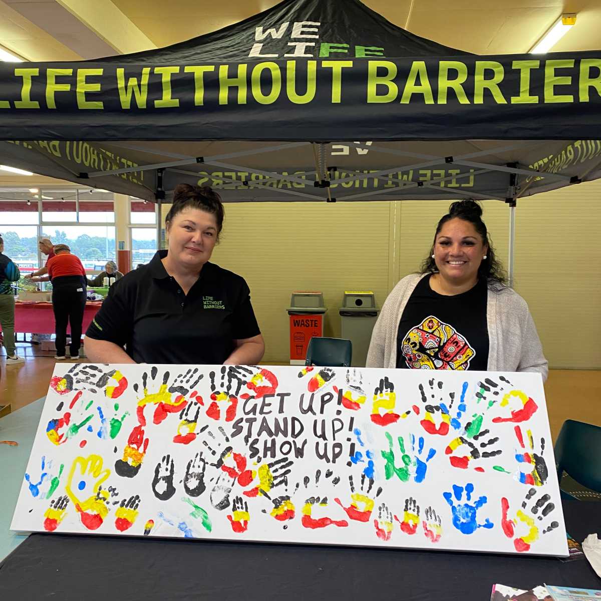 Two women stand under a Life Without Barriers banner, holding up an artwork with hand prints and text that reads "Get Up, Stand Up, Show Up"