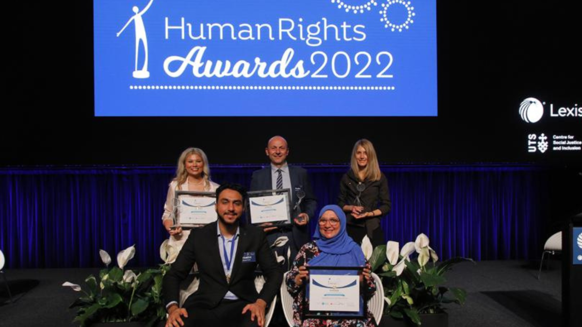 Five recipients of the Human Rights Awards standing together as a group in front of a stage, smiling at the camera.