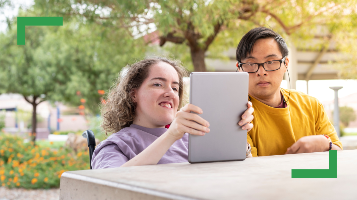 A woman in a purple T-shirt sitting at an outdoor table holding a computer tablet. Sitting next to her is a man wearing glasses and a yellow shirt who is also looking at the tablet.