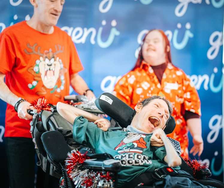 A participant wears a green Christmas shirt and smiles. Behind him is a support worker wearing a red Christmas shirt.