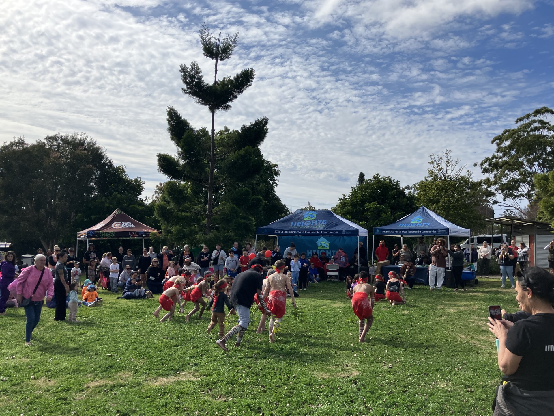 A group of young people in traditional body paint and red clothing performing in front of a crowd. 