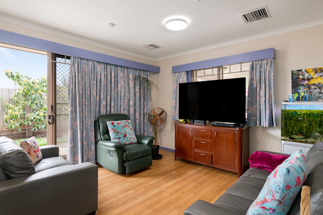 Cosy living room with wooden flooring, two grey leather sofas and a green recliner chair, all with floral cushions. A large flat-screen TV sits on a wooden cabinet, with light blue patterned curtains covering the windows and sliding door. An indoor plant and a fish tank with lush greenery add a natural touch to the space.
