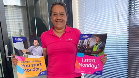 Roylene is standing in an office wearing a pink Joblife shirt. She is holding two brochures. Ones says 'you start Monday' the other says 'I start Monday'.