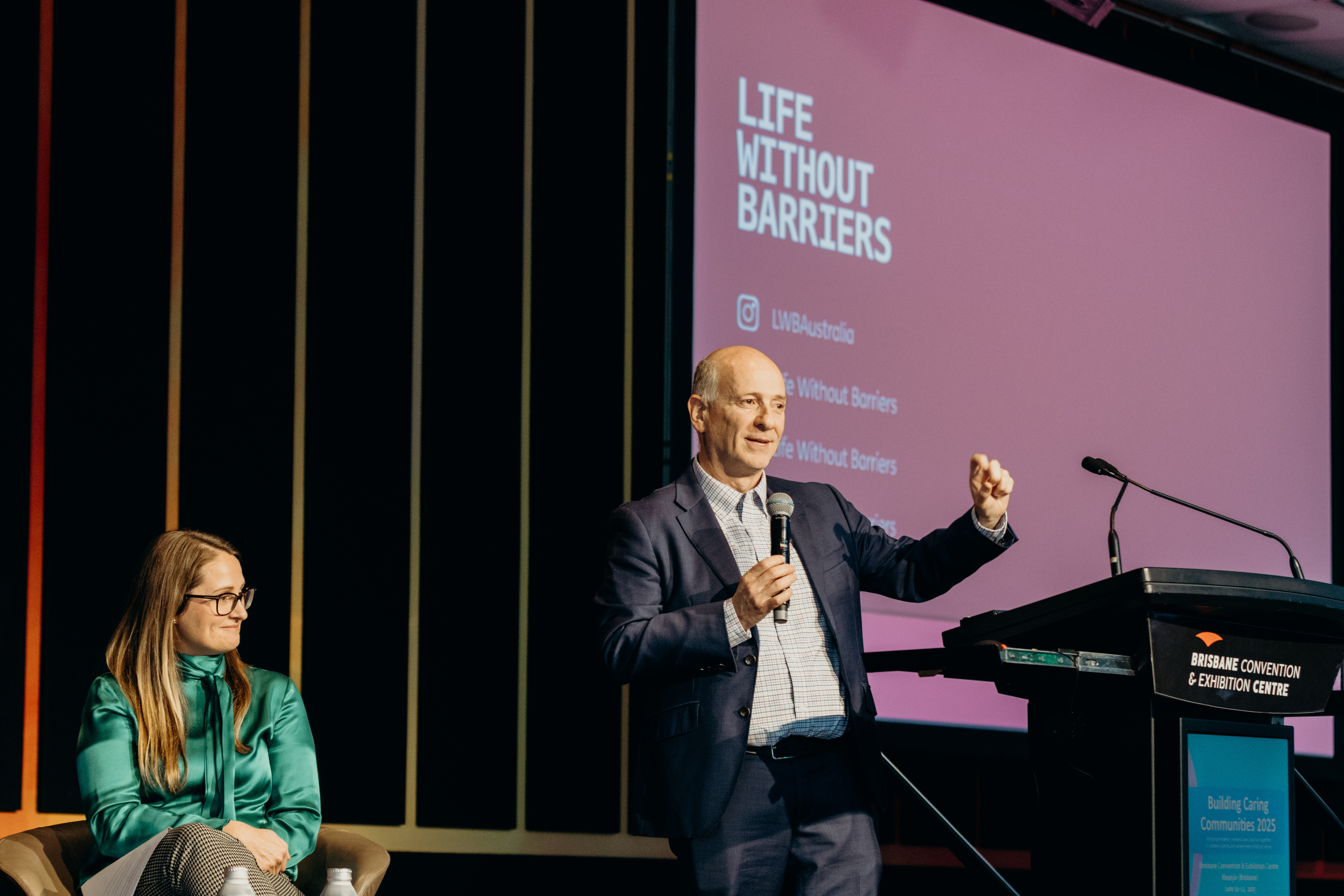 Anthony and Carly are on stage. Carly is sitting and Anthony is standing and holding a microphone. There is a pink screen with 'Life Without Barriers' in the background.