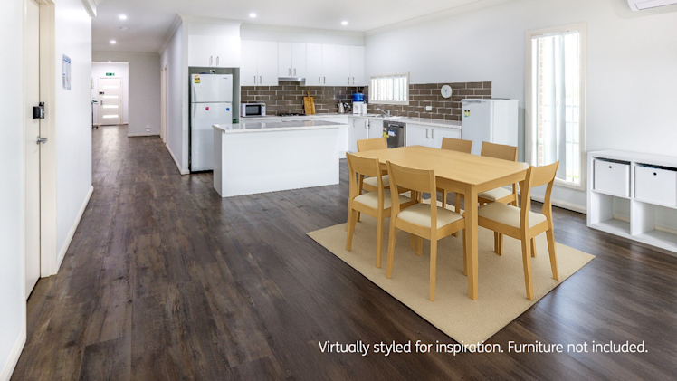 Open-plan kitchen and dining area featuring a white island bench, wooden dining table with six chairs, dark wood flooring, and white cabinetry with a brown tile backsplash.