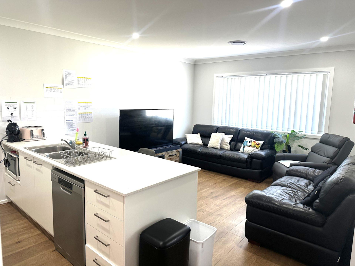 Cozy living area with three black leather lounges, a large TV on a wooden entertainment unit, indoor plant, and wide window with vertical blinds.