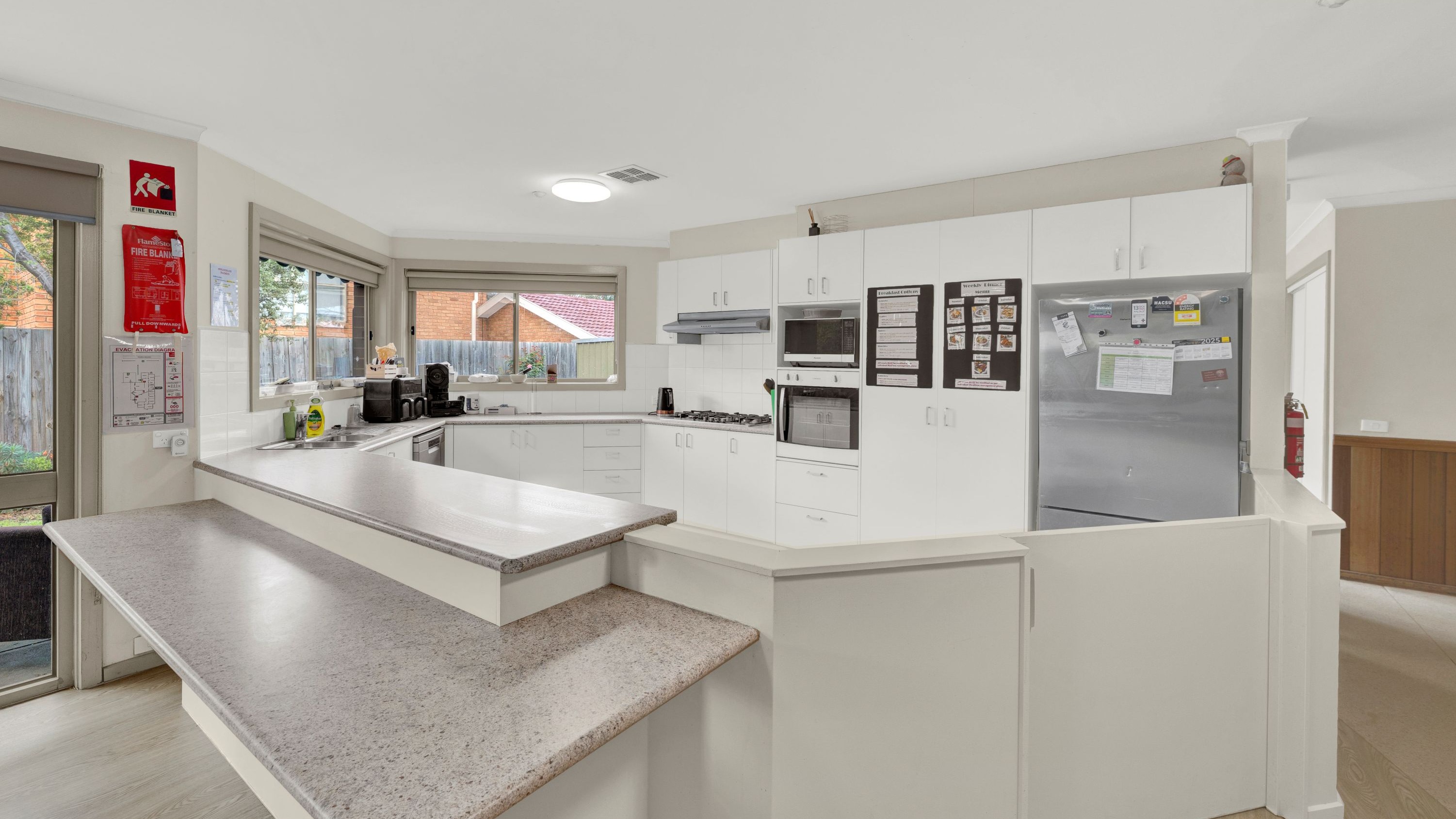 Accessible shared kitchen with white cabinetry, bench seating, built-in oven, cooktop, and wide walkways.