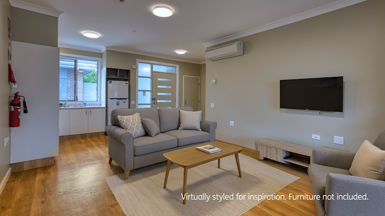 Open-plan living space with a grey sofa set, mounted TV, light wood furniture, and partial view of the kitchen and front door.