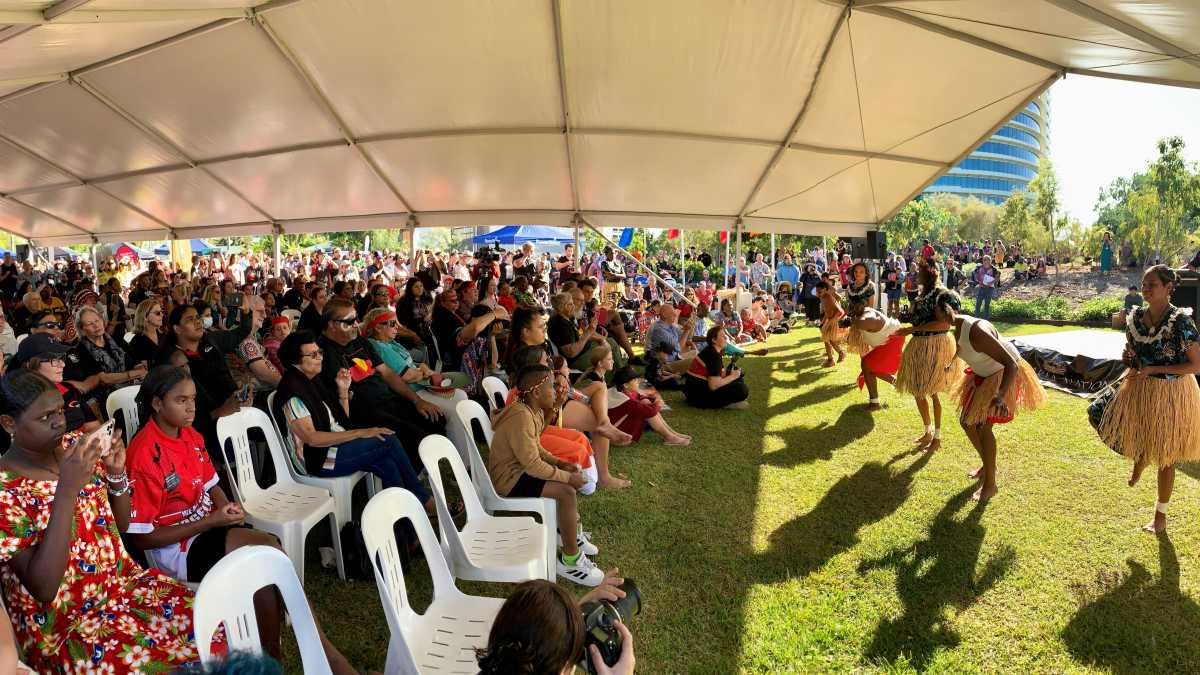A crowd sits under a pavilion watching a group of Aboriginal women dancing.