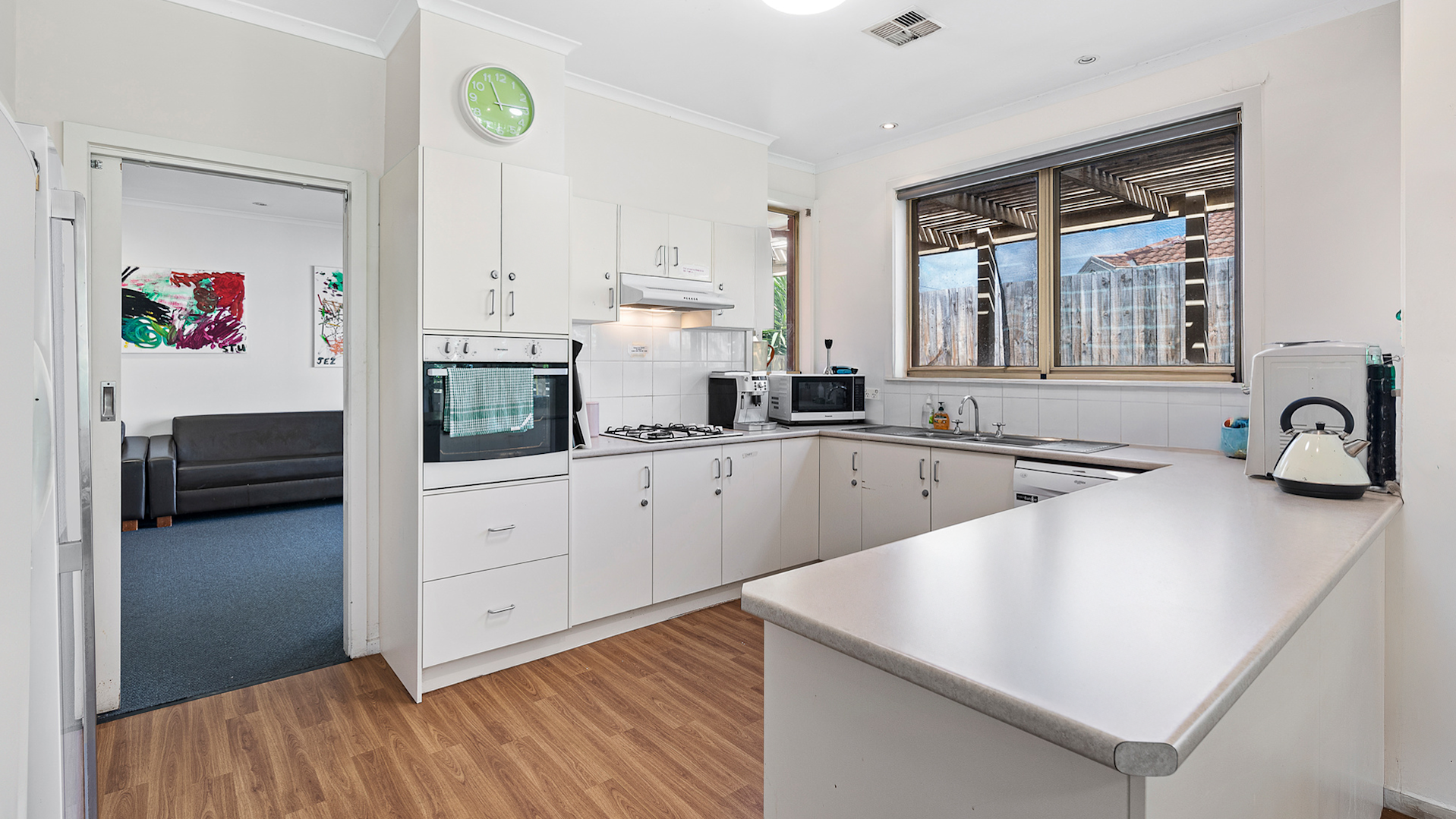 U-shaped kitchen with white cabinets and counters, wooden floorboards and a window looking out to the backyard.
