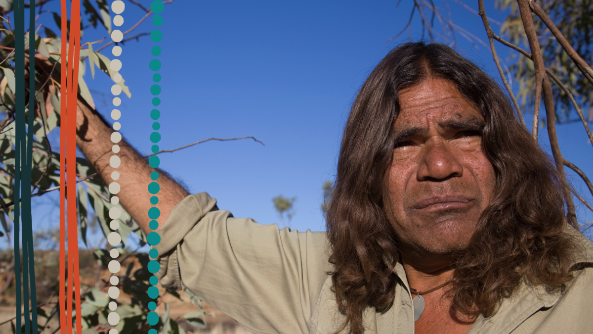 An Aboriginal man with shoulder-length brown hair is wearing a beige shirt whilst standing underneath a gum tree.