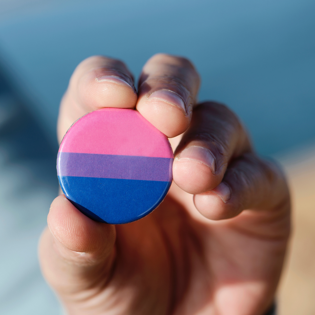 Close-up of a hand holding a pin with the bisexual flag colours on it.