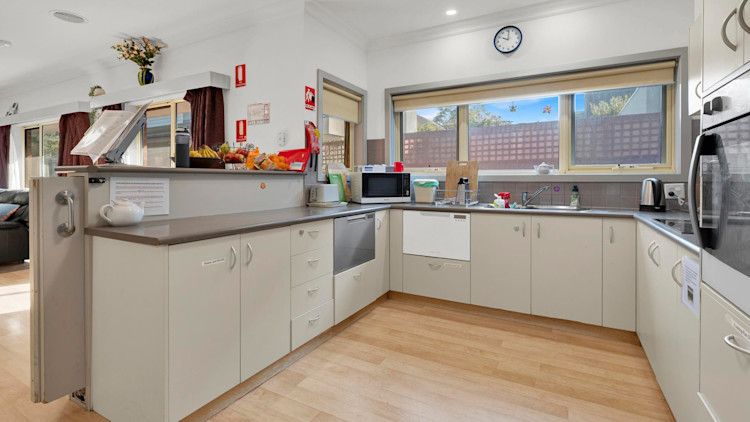 Functional kitchen with cream cabinetry, under-bench appliances, a large window providing natural light, and various kitchen items neatly arranged on countertops.