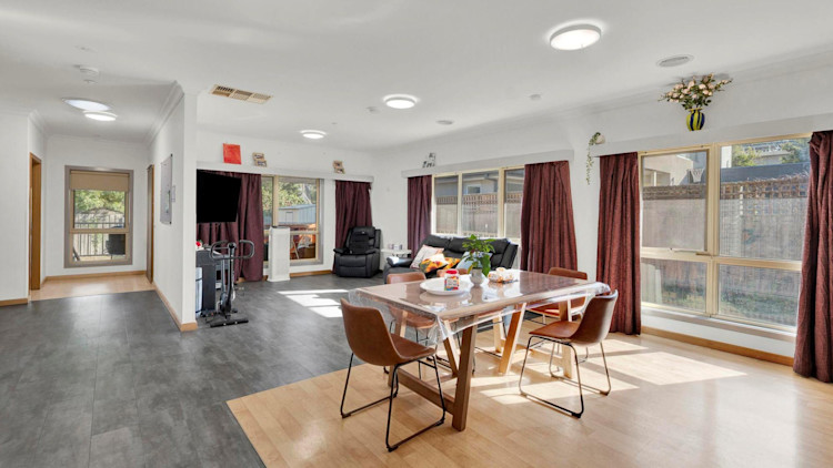 Spacious, sunlit communal living and dining area in a residential facility, featuring a dining table with chairs, lounge seating, exercise equipment, and large windows with maroon curtains. The room has a combination of wood and dark tile flooring and is decorated with minimal ornaments.