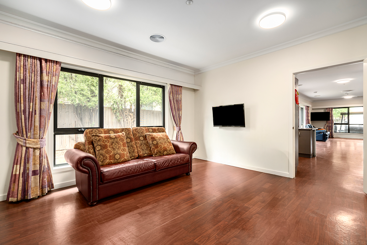 Living area with a maroon leather sofa and patterned cushions, wall-mounted TV, large window with purple curtains, and view into adjoining open-plan room.