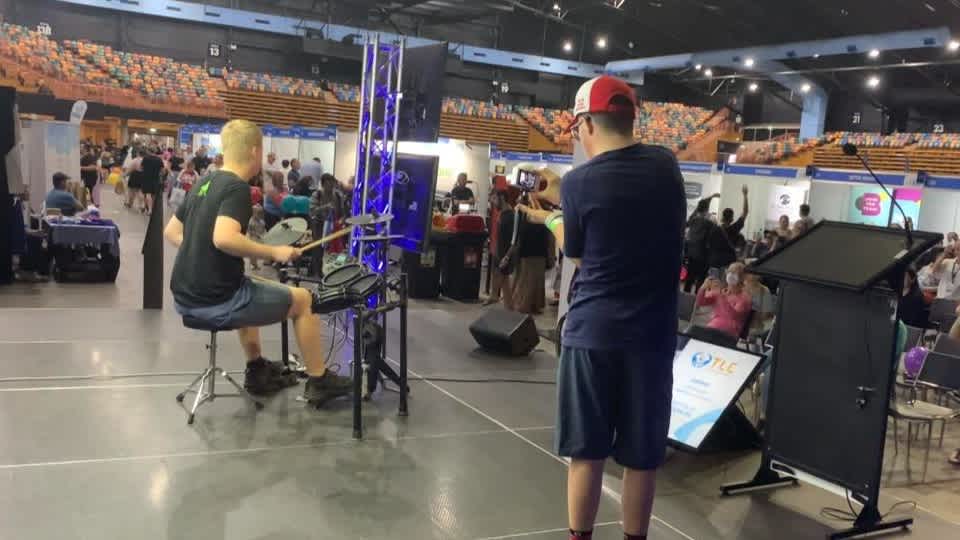 Alex is playing drums on the stage in front of a crowd. Patrick stands next to him holding a camera.