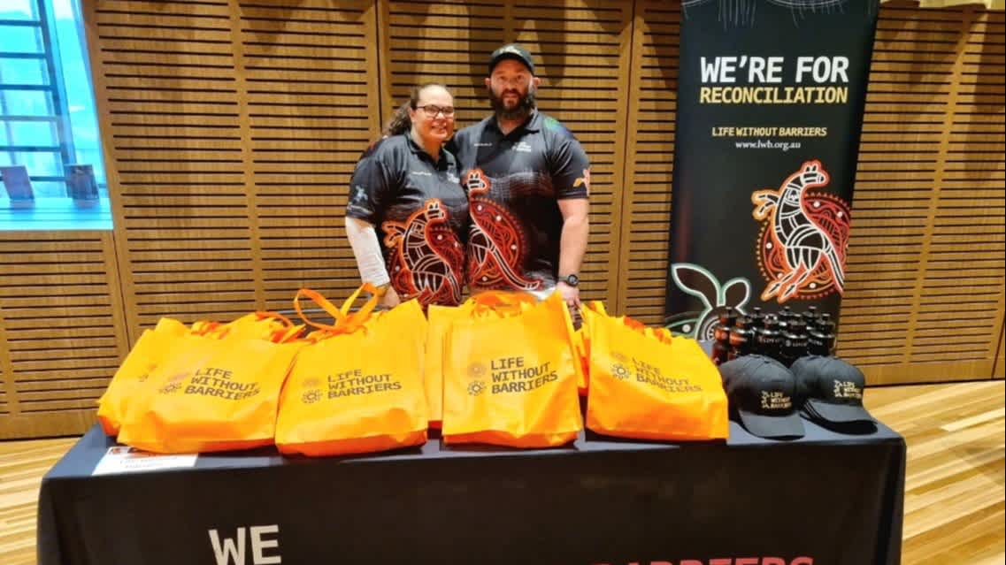 Adam Towney and Gayle wearing black polos with an Aboriginal design on them, standing at a Life Without Barriers stall.