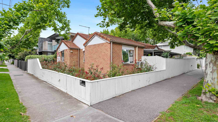 Front view of a single-storey brick home with a white fence, garden beds and mature street trees.