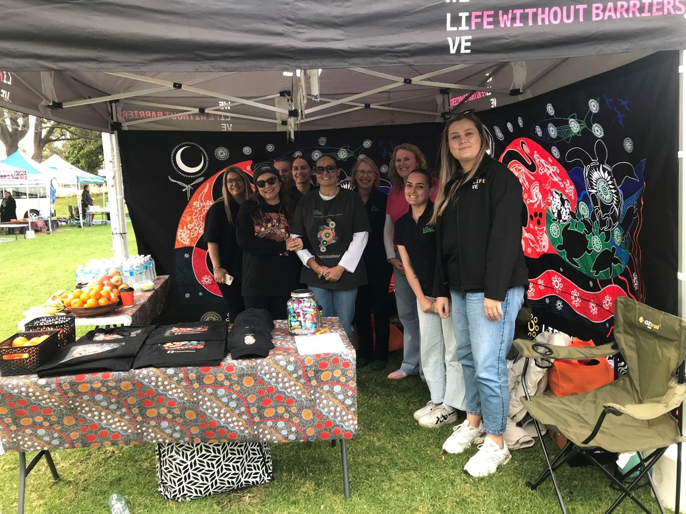 A group of staff at the Warrawong NAIDOC Family Fun Day, standing behind the Life Without Barriers stall and smiling at the camera.