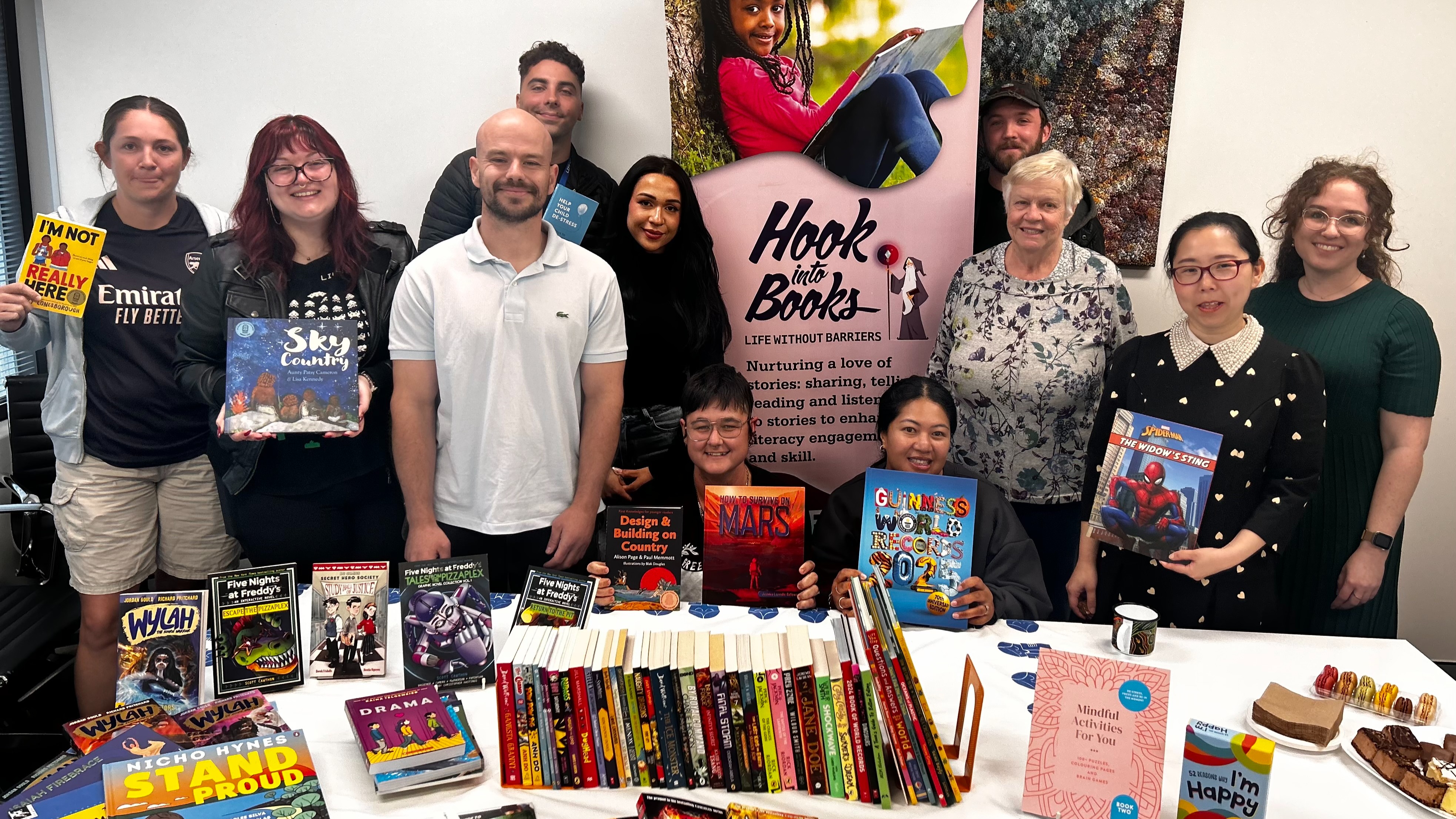 A group of staff stand behind a table covered in books, smiling at the camera. Some staff members hold books in their hands. Behind them is the Hook into Books™ banner.