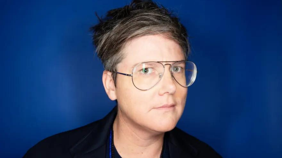 Hannah Gadsby standing in front of a blue background wearing black clothing and glasses.