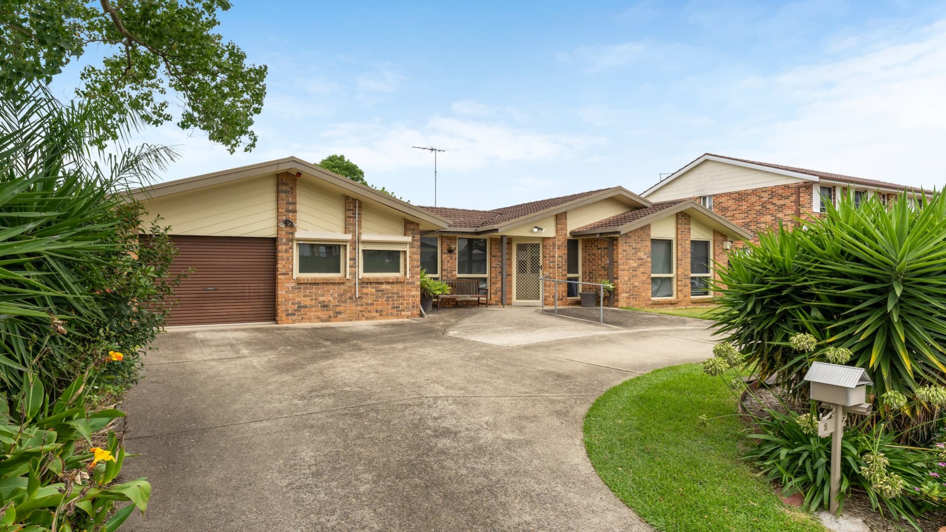 House with a brick facade and a concrete driveway with grass and shrubs on either side leads to the garage and the front door.