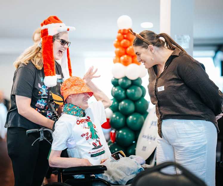 A participant wearing a white Christmas shirt and red hat laughs with two support workers.