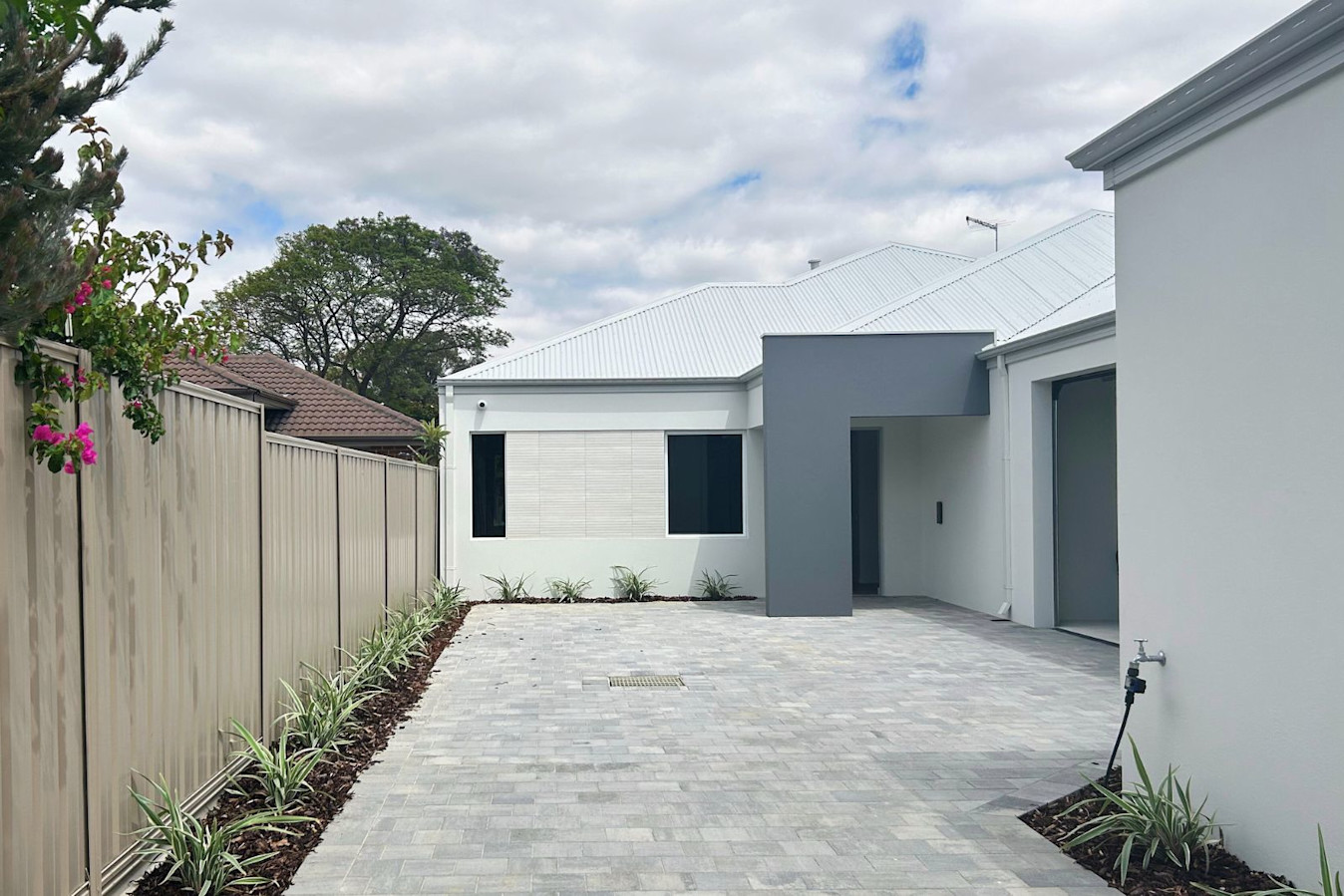 Modern single-storey home with light grey roof, paved driveway, and landscaped garden beds along a beige fence.