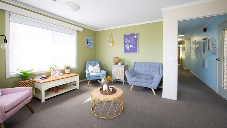 Bright living room with green walls, blue and pink chairs, artwork, and a round wicker coffee table.