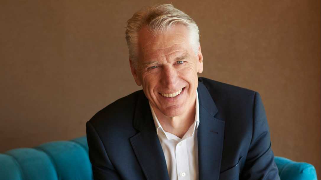Greg Ridder is wearing a white shirt and dark blue suit. He has white hair and is smiling into the camera. He has his hands folded together and is sitting on a blue armchair. The wall behind him in a bronze colour.