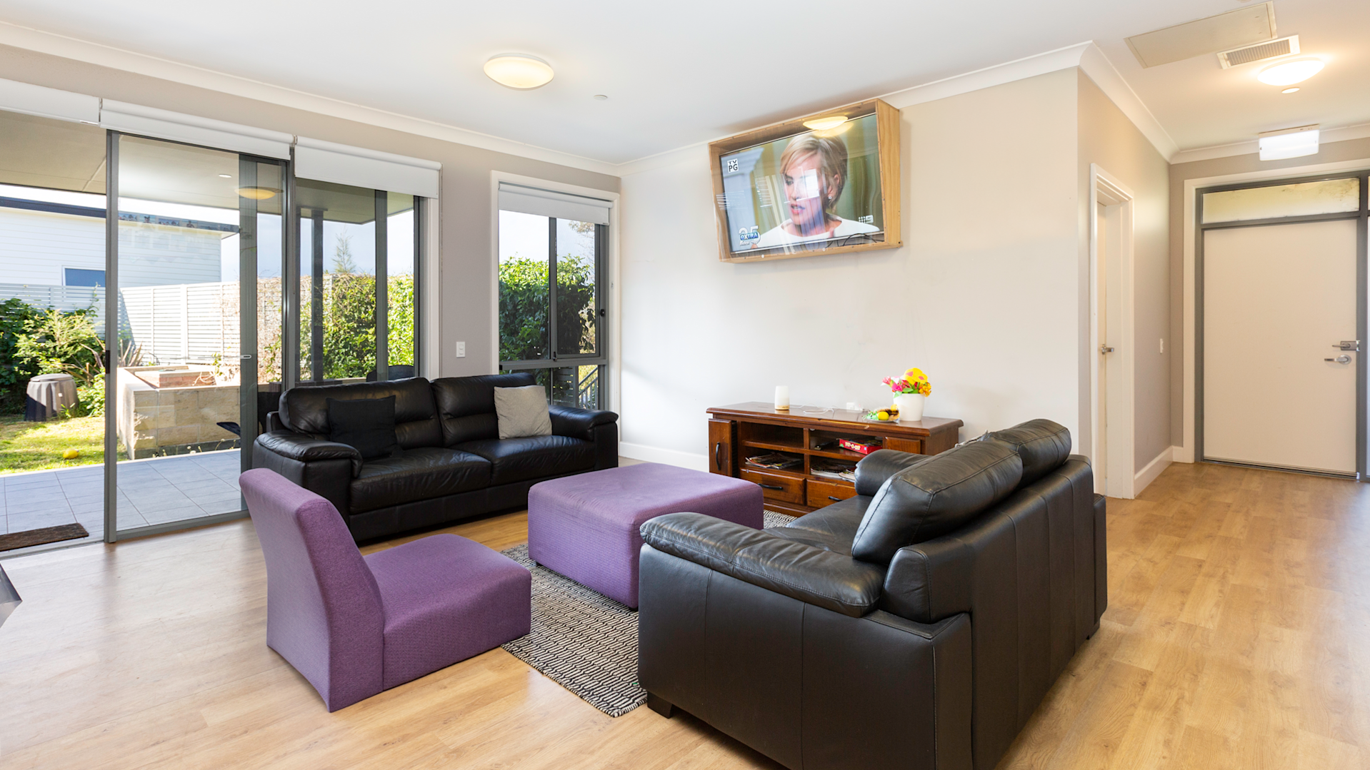Comfortable living room with black leather sofas, purple ottomans, wall-mounted TV, and large glass doors opening to a garden patio.