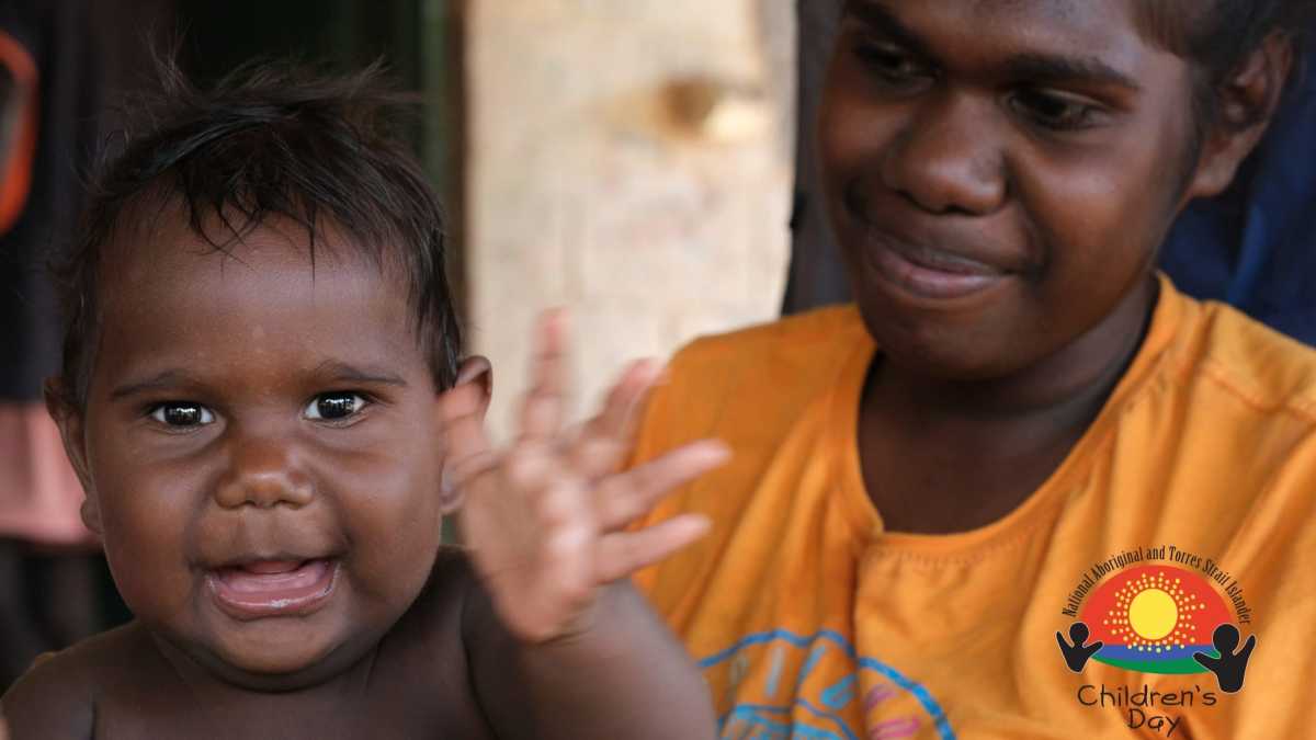 An Aboriginal toddler is smiling and waving at the camera. Behind the child is an Aboriginal woman wearing an orange T-Shirt who is looking at the child and smiling.