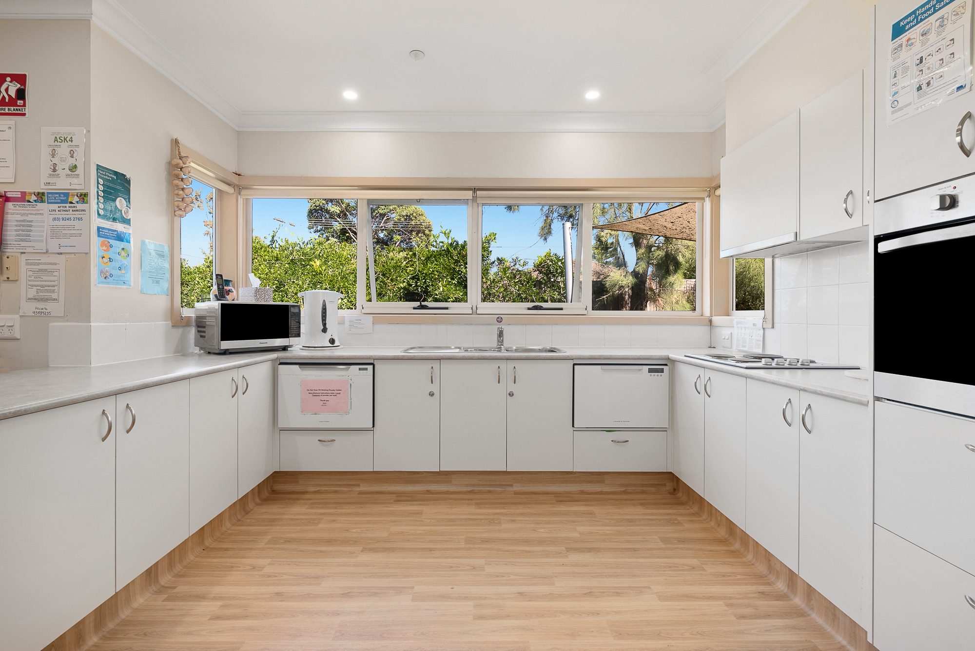 Bright accessible kitchen with large windows, double sink, and white cupboards.