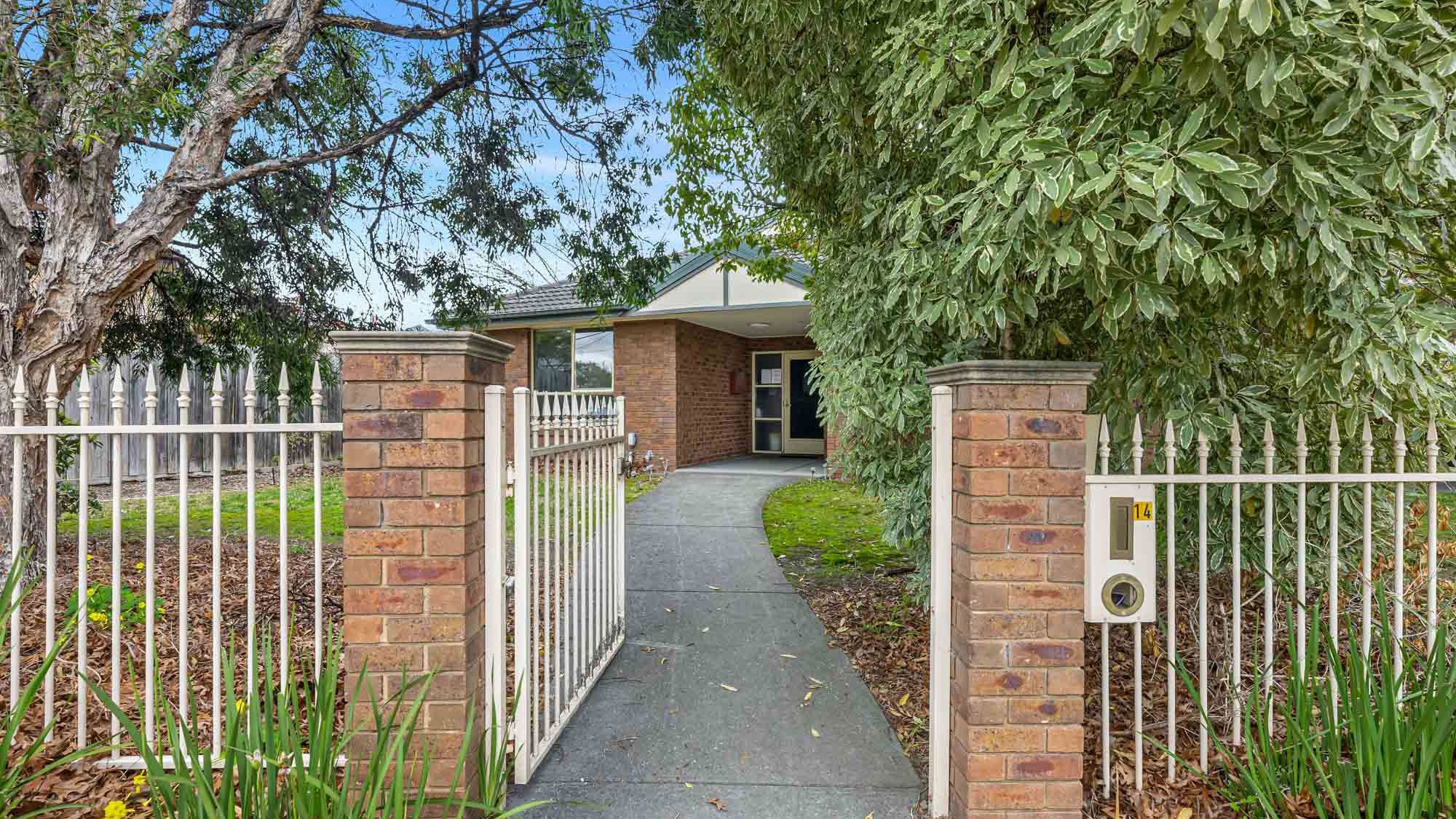 Pathway leading through a gated fence to the main entrance, framed by mature trees and garden beds.
