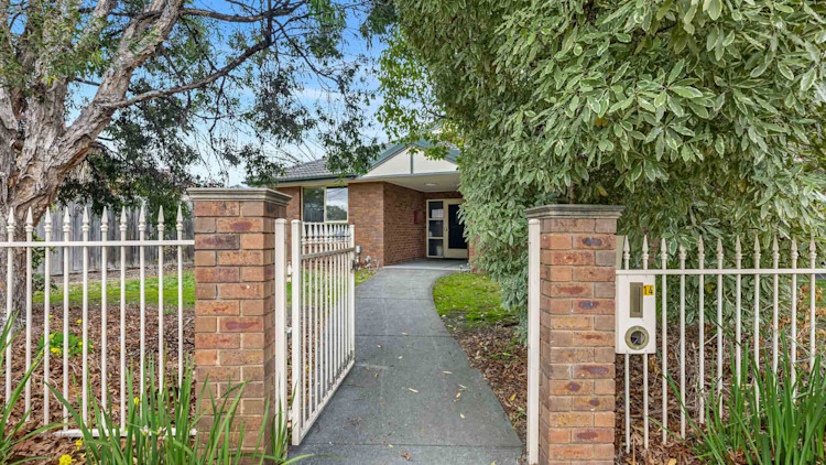 Pathway leading through a gated fence to the main entrance, framed by mature trees and garden beds.