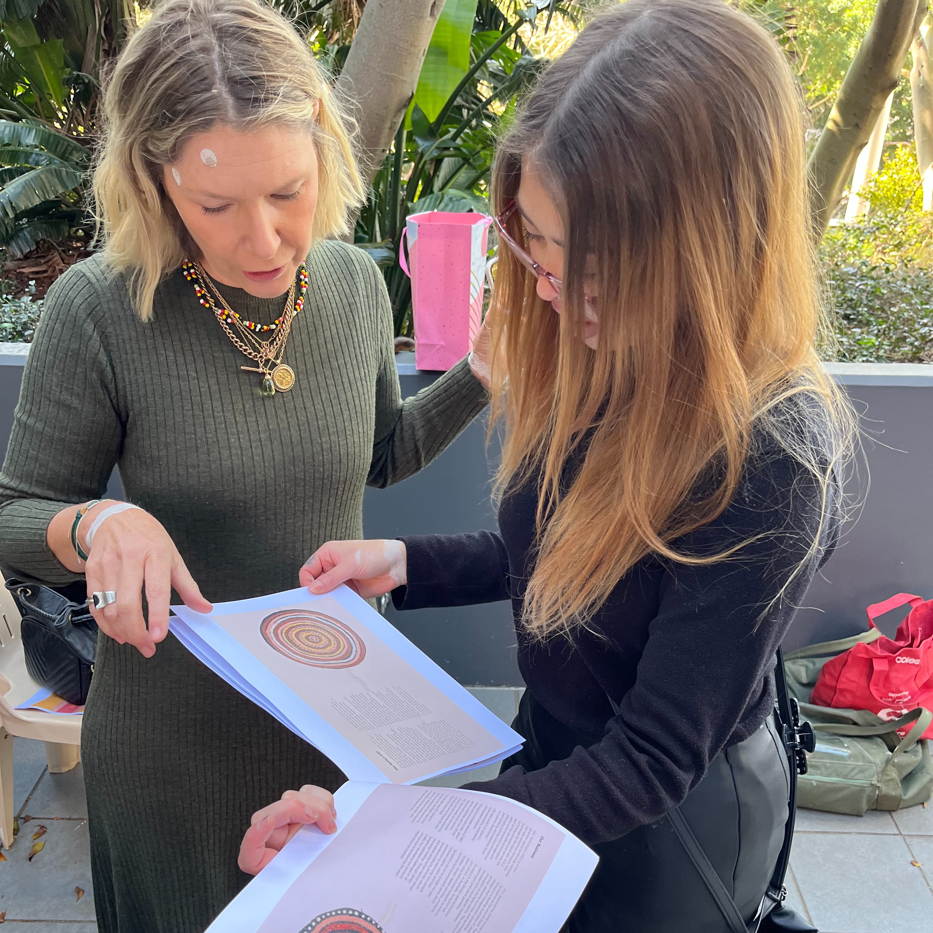 Shannon and a woman with blonde hair standing next to each other, looking at a booklet with Aboriginal artwork.