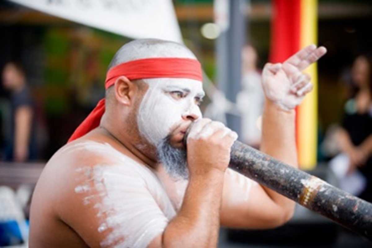 Tyne Smith is playing the didgeridoo and wearing white face and body paint and a red headband.