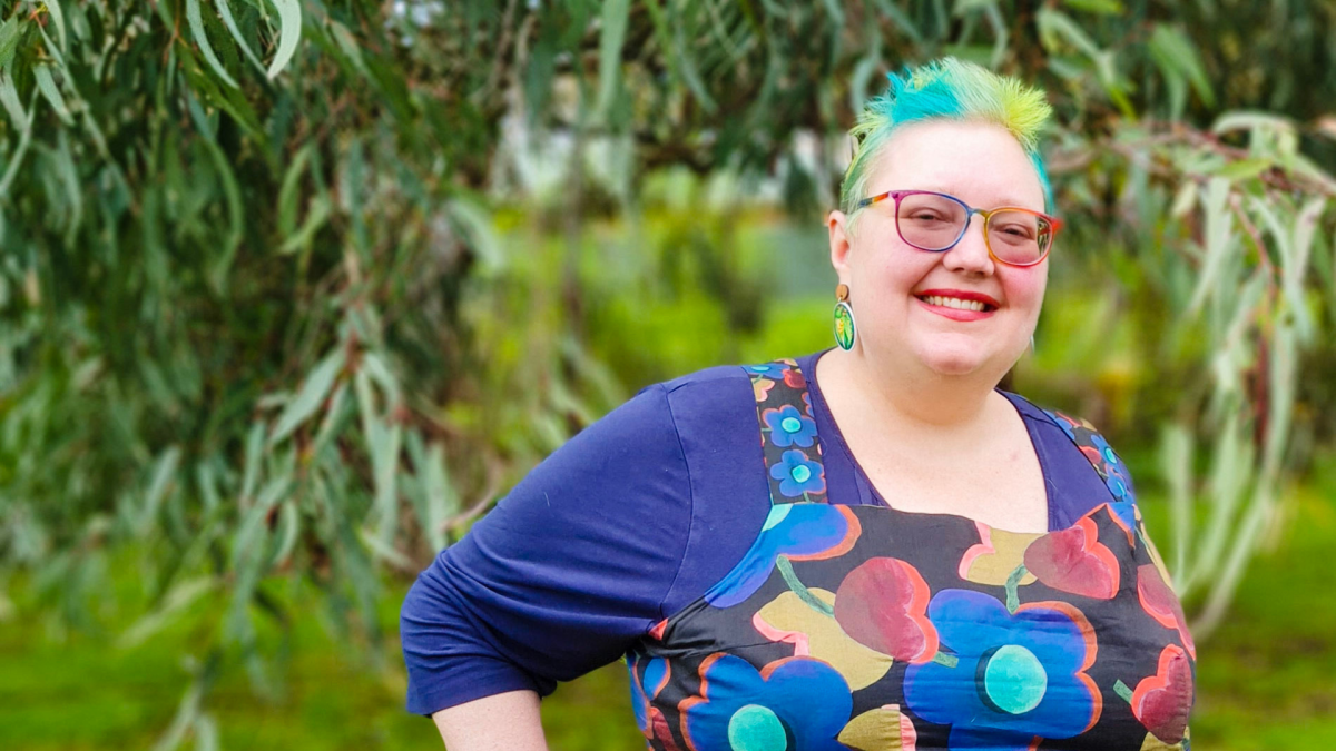 Ceara standing outdoors wearing a long-sleeved purple top under a dress with blue, yellow and red flowers. Ceara has short, colourful hair and rainbow glasses.