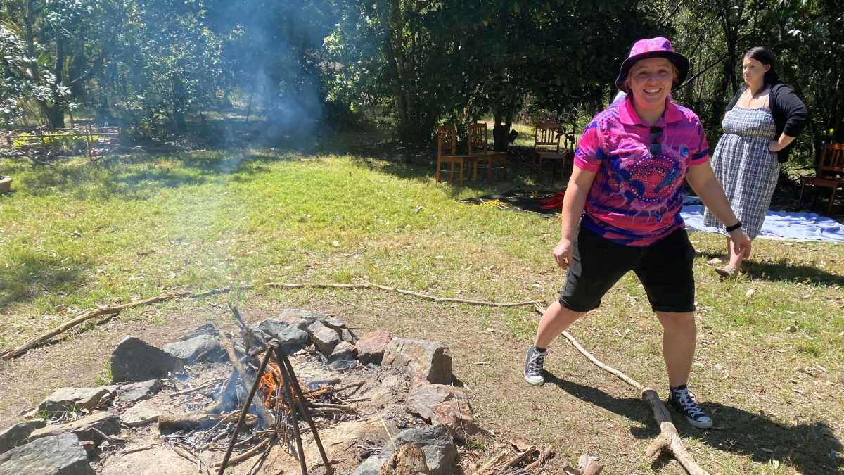 A woman wearing black shorts, a pink top and a pink hat stands next to a camp fire smiling at the camera.