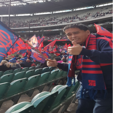 Ralph is wearing a Melbourne Football Club scarf and jumper. He is standing at the MCG looking at the camera with his thumbs up.