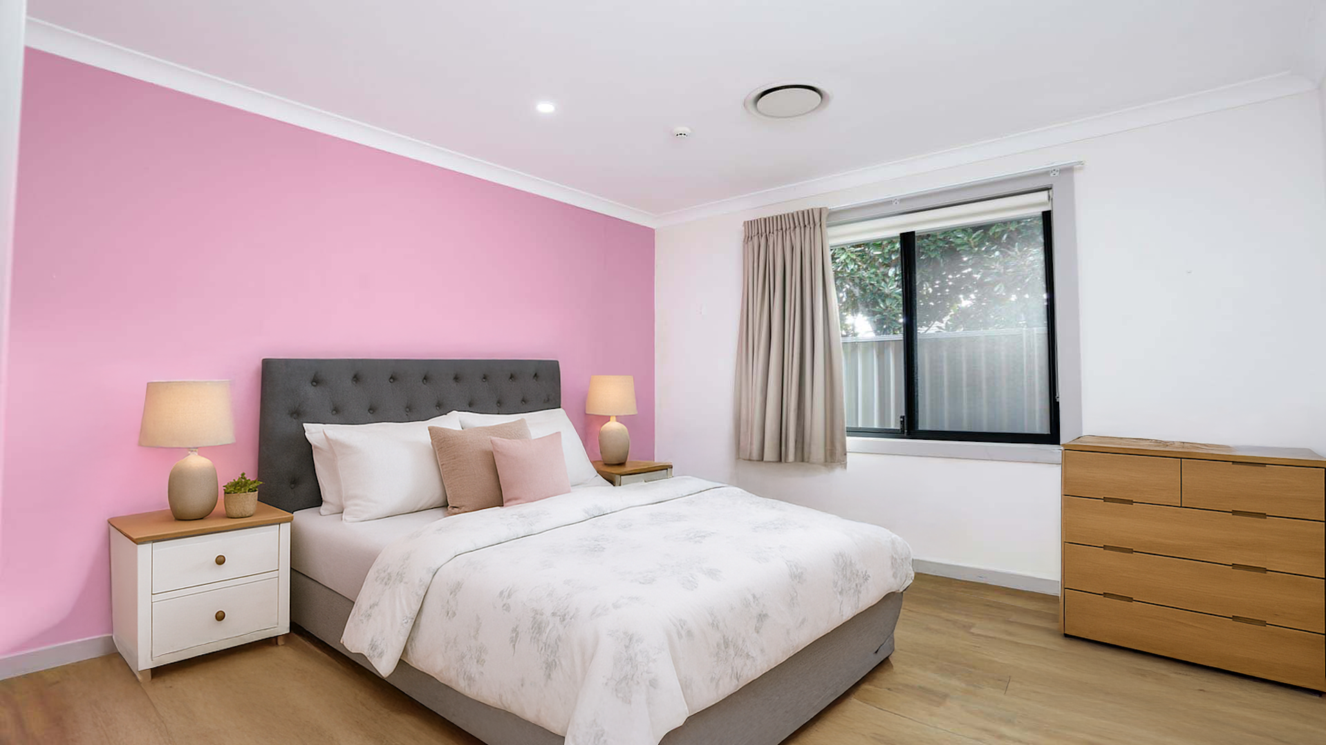 Modern bedroom with a pink feature wall behind a grey upholstered bed, white bedding, neutral-toned pillows, matching bedside tables with lamps, a wooden dresser, and a large window with beige curtains.