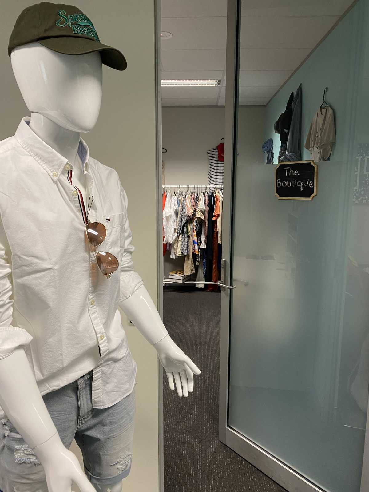 A mannequin wearing a cap, white shirt and denim shorts next to a door with a sign that reads 'The Boutique'.