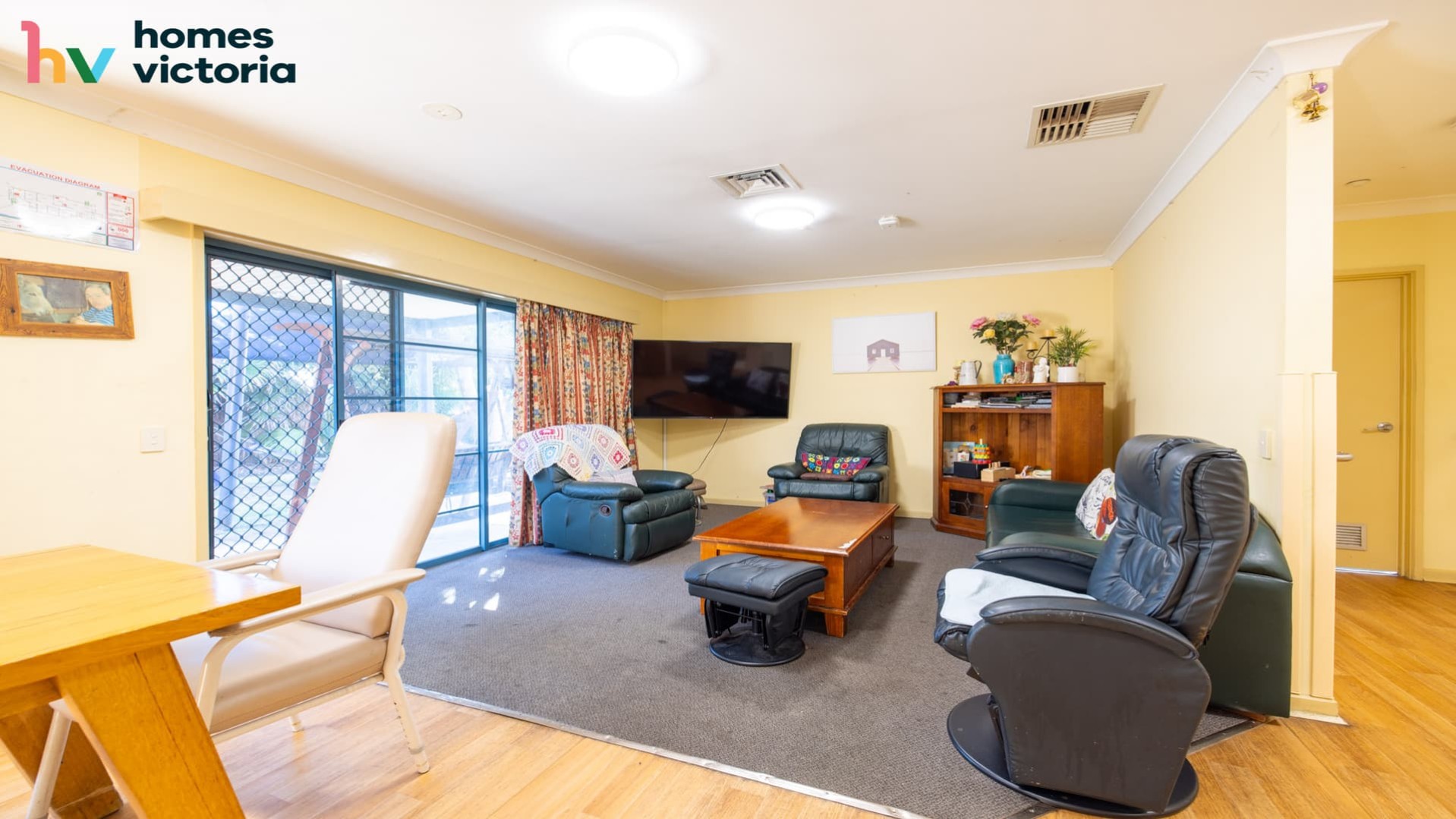 Spacious loungeroom with easy chairs and couch on grey carpet facing wall mounted TV.  Room has sliding door to access backyard.